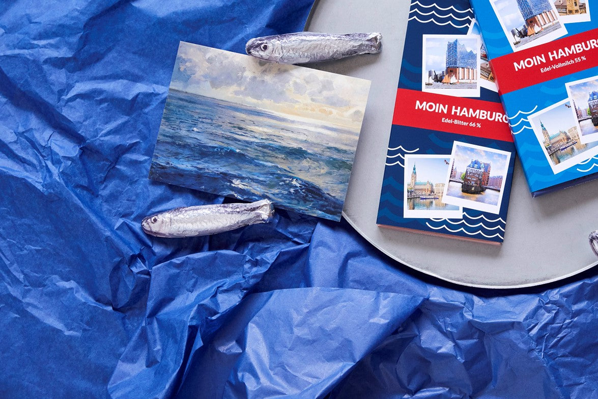 Zwei Schokoladentafeln im Hamburg Branding und Vollmilchschokoladen in Sardinen-Optik sind auf einem blauen Hintergrund plaziert.