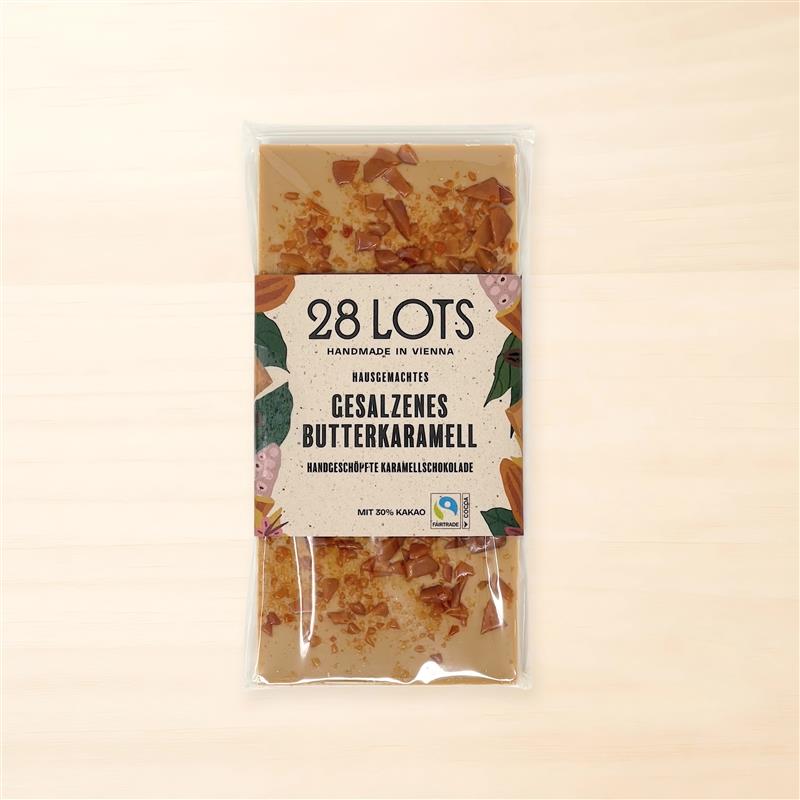 Schokoladentafel in transparenter Folienhülle mit mittiger Papierbanderole: cremefarben, fein gesprenkelt, mit bunten Pflanzen-/Blattillustrationen an den Rändern. Vorn groß „28 LOTS“, darunter „Handmade in Vienna“ und „Gesalzenes Butterkaramell“, unten Fairtrade-Kakao-Siegel. Durch die Folie sieht man helle Karamellschokolade mit karamelligen Stückchen.