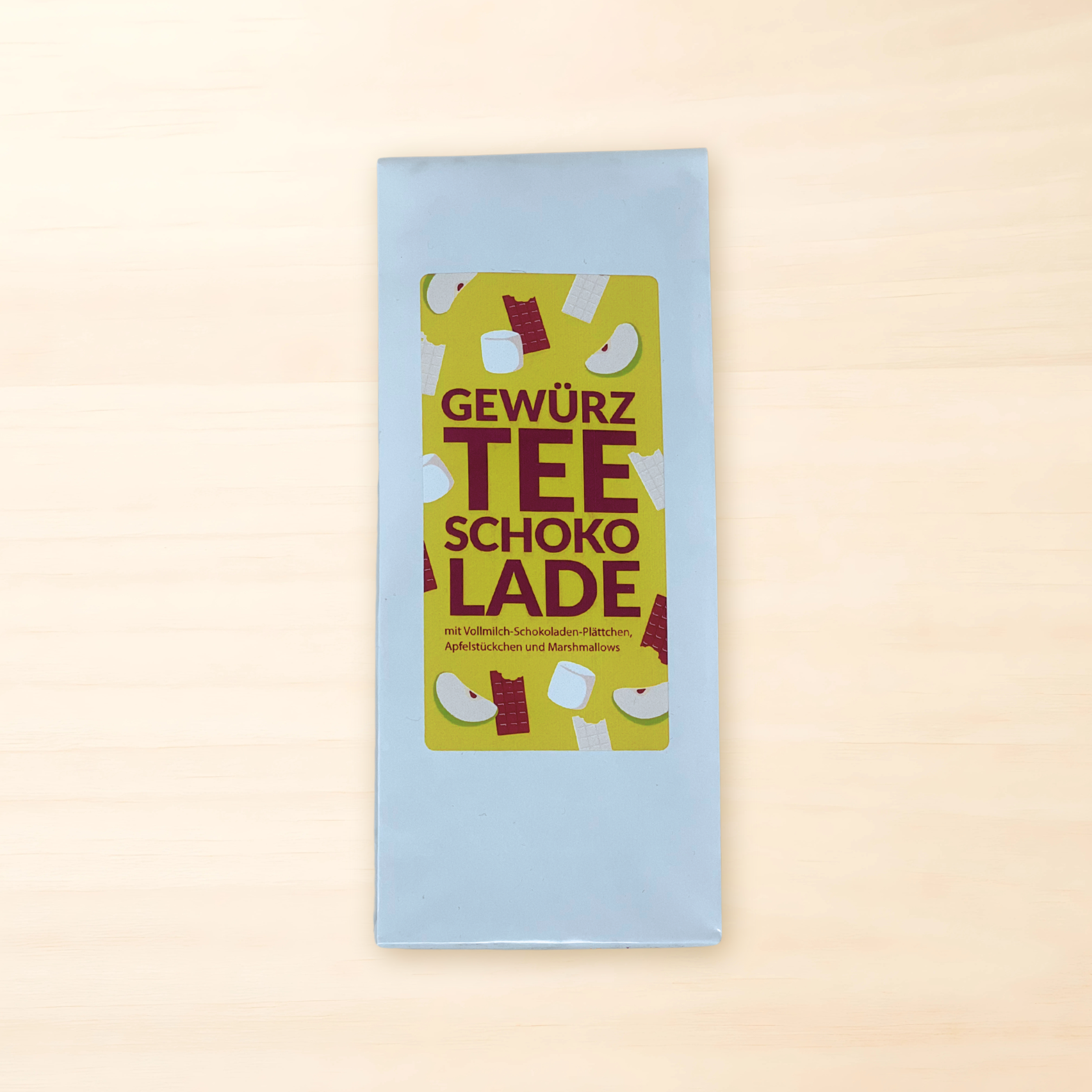 „Weiße Teeverpackung mit gelbem Etikett, darauf Illustrationen von Schokolade, Apfelstücken und Marshmallows sowie der Aufschrift ‚Gewürz Tee Schokolade‘.“