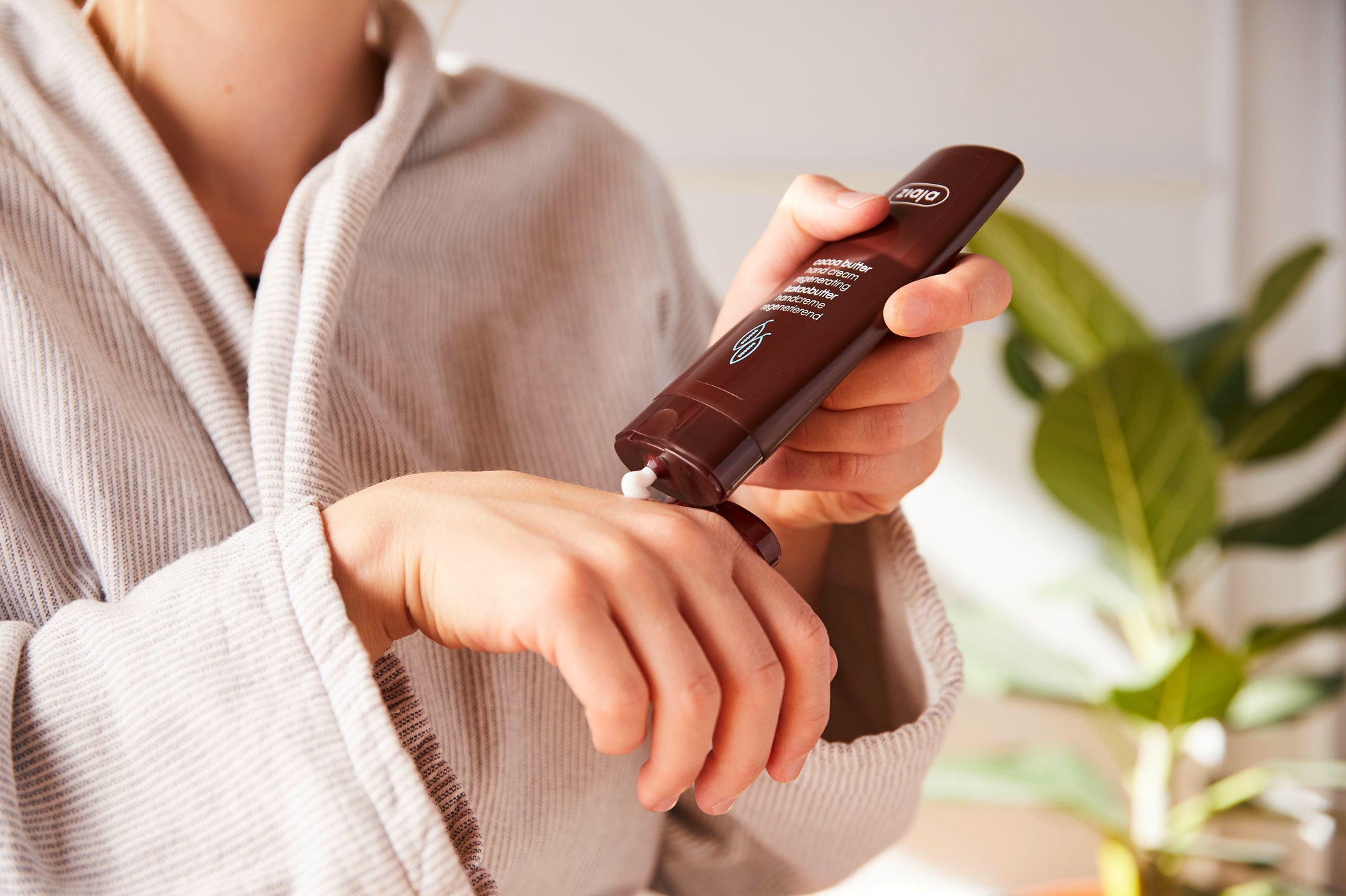 Im Vordergrund sieht man eine Hand, auf die gerade Creme der Marke Ziaja aufgetragen wird. Dazu eine Person im Bademantel und im Hintergrund eine grüne Pflanze.