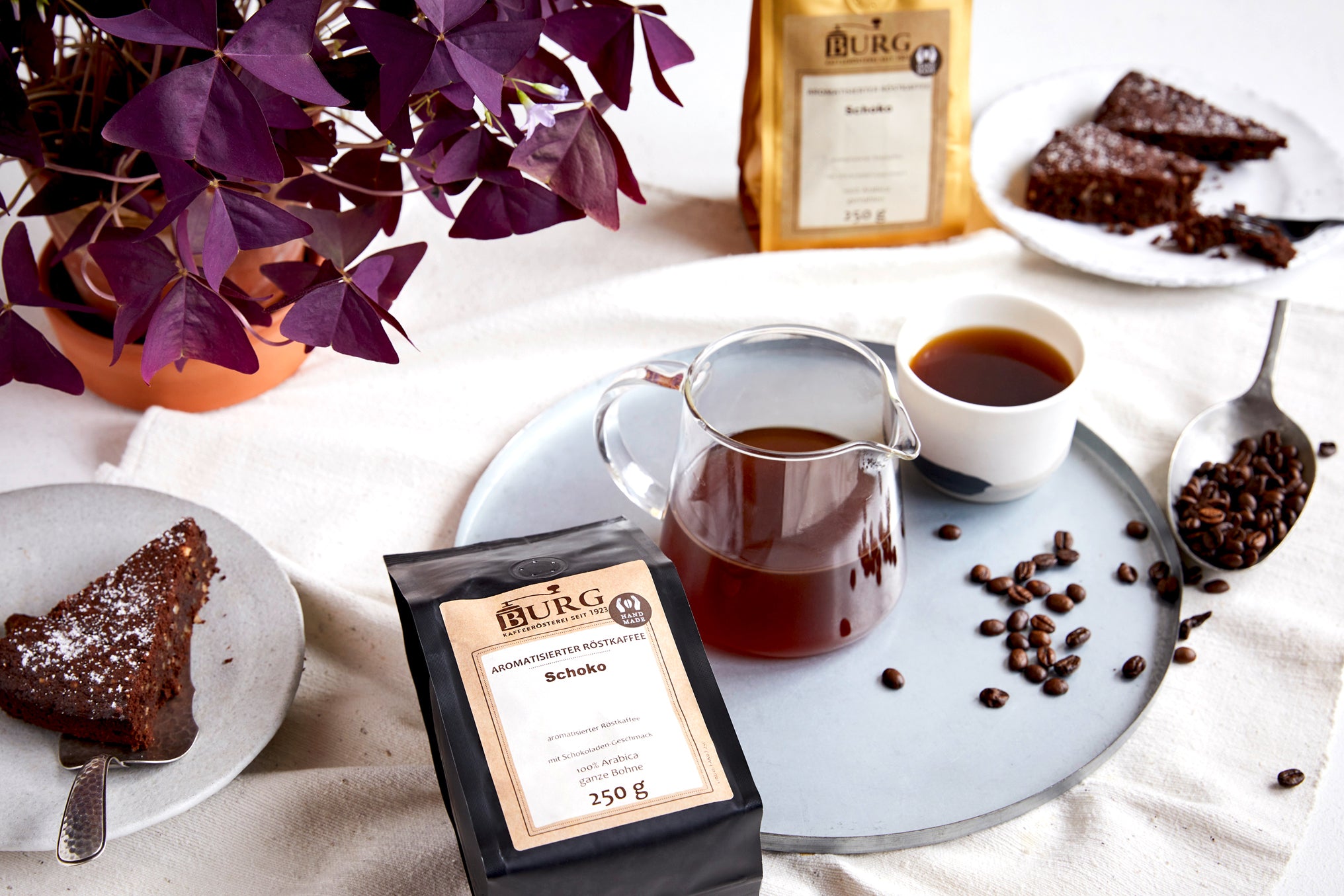 Kaffeetafel mit Schokoladenkuchen, Tasse Filterkaffee, Kaffeebohnen und einer Packung BURG Schoko-Röstkaffee.