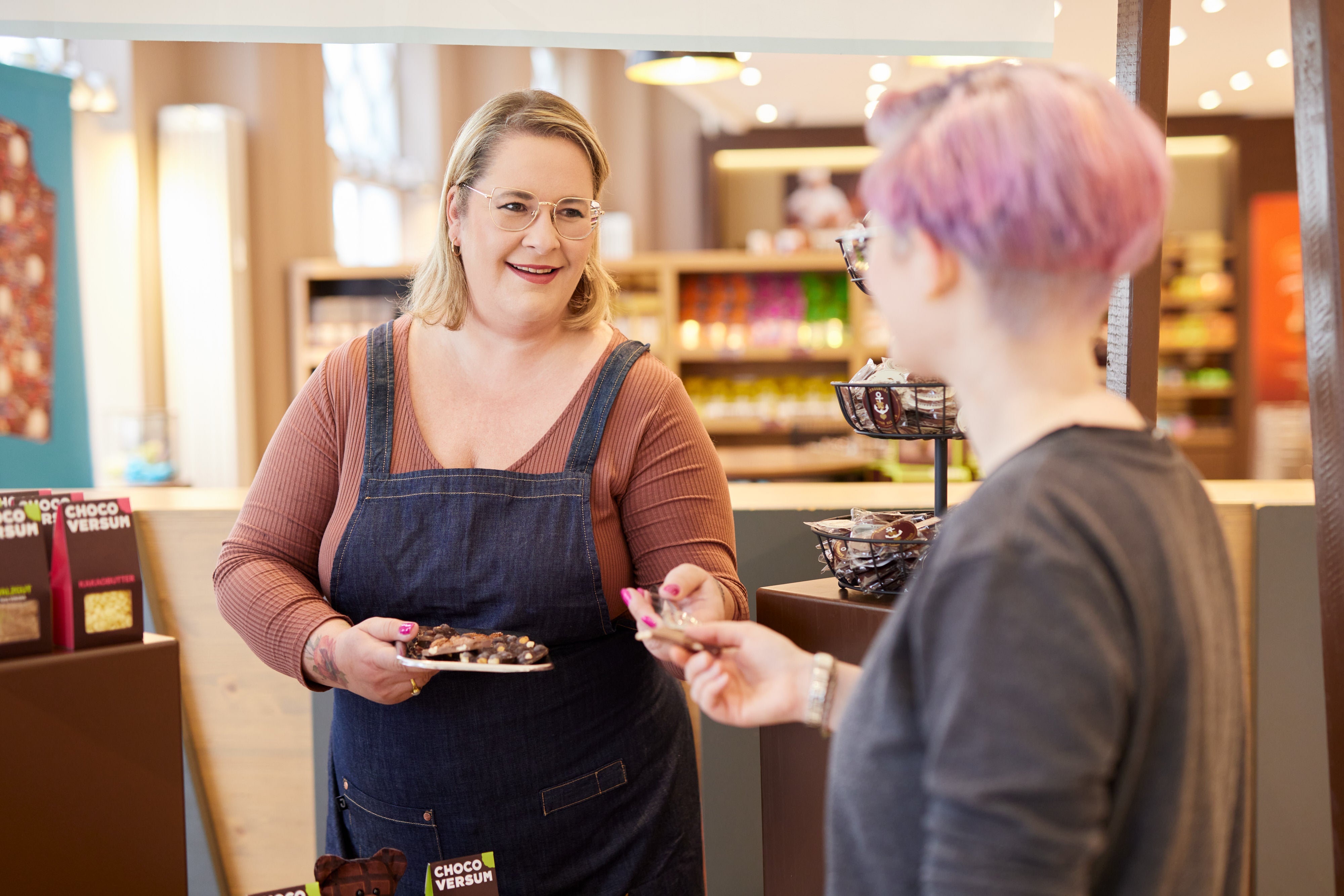 Frau mit Schürze bietet im Schokoladen-Shop einem Gast Schokoladenproben an.