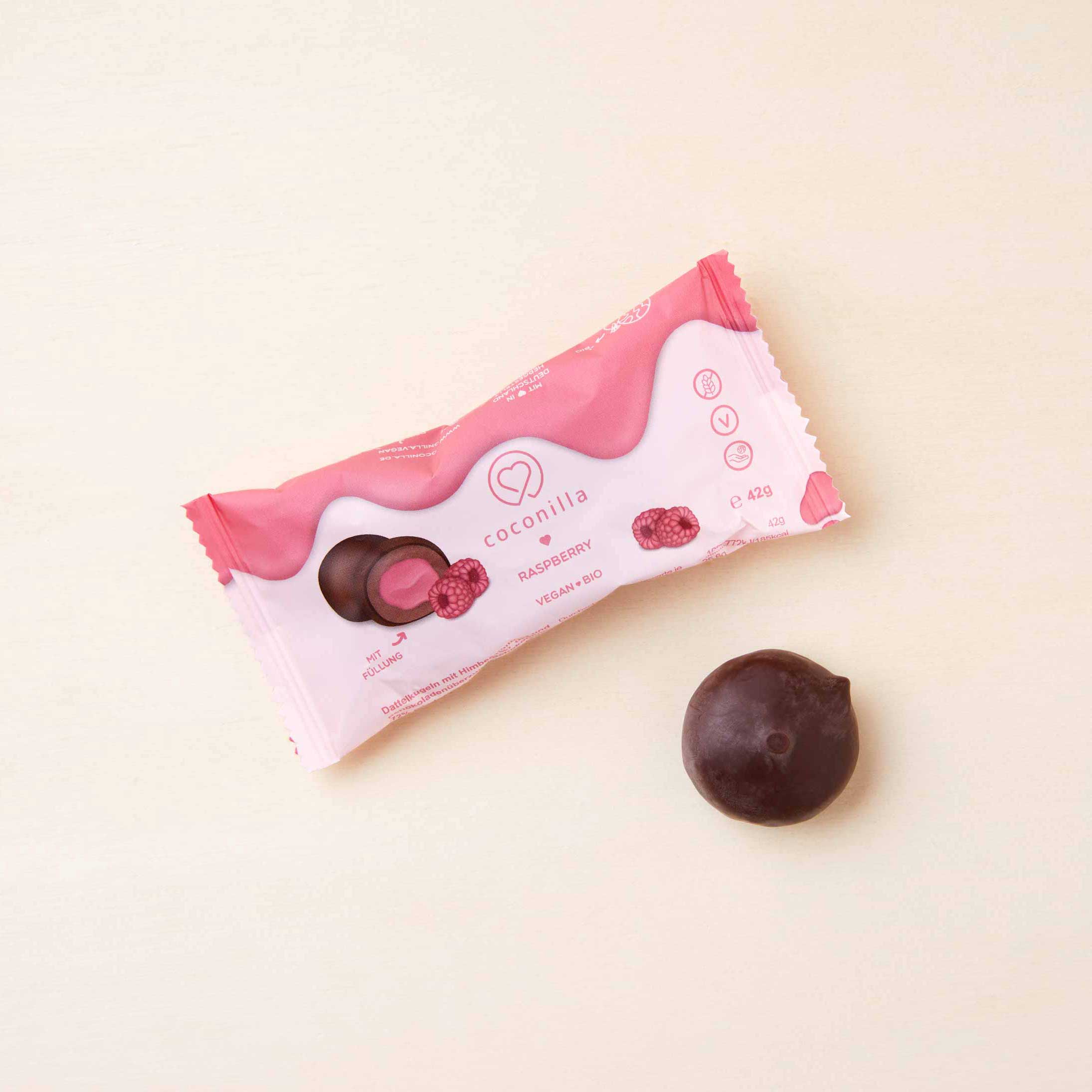 rosafarbene Plastikverpackung und eine ausgepackte Dattelkugel mit Himbeerfüllung von Coconilla