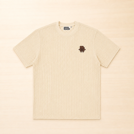 Cremefarbenes Kinder-T-Shirt mit Waffelstruktur und kleiner aufgestickter Schokoladenfigur auf der linken Brust.