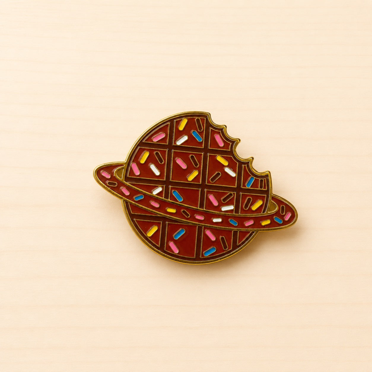 Runder Metall-Ansteck-Pin als brauner Schokoladenplanet mit bunten Streuseln, umlaufendem Ring und goldfarbenen Konturen.