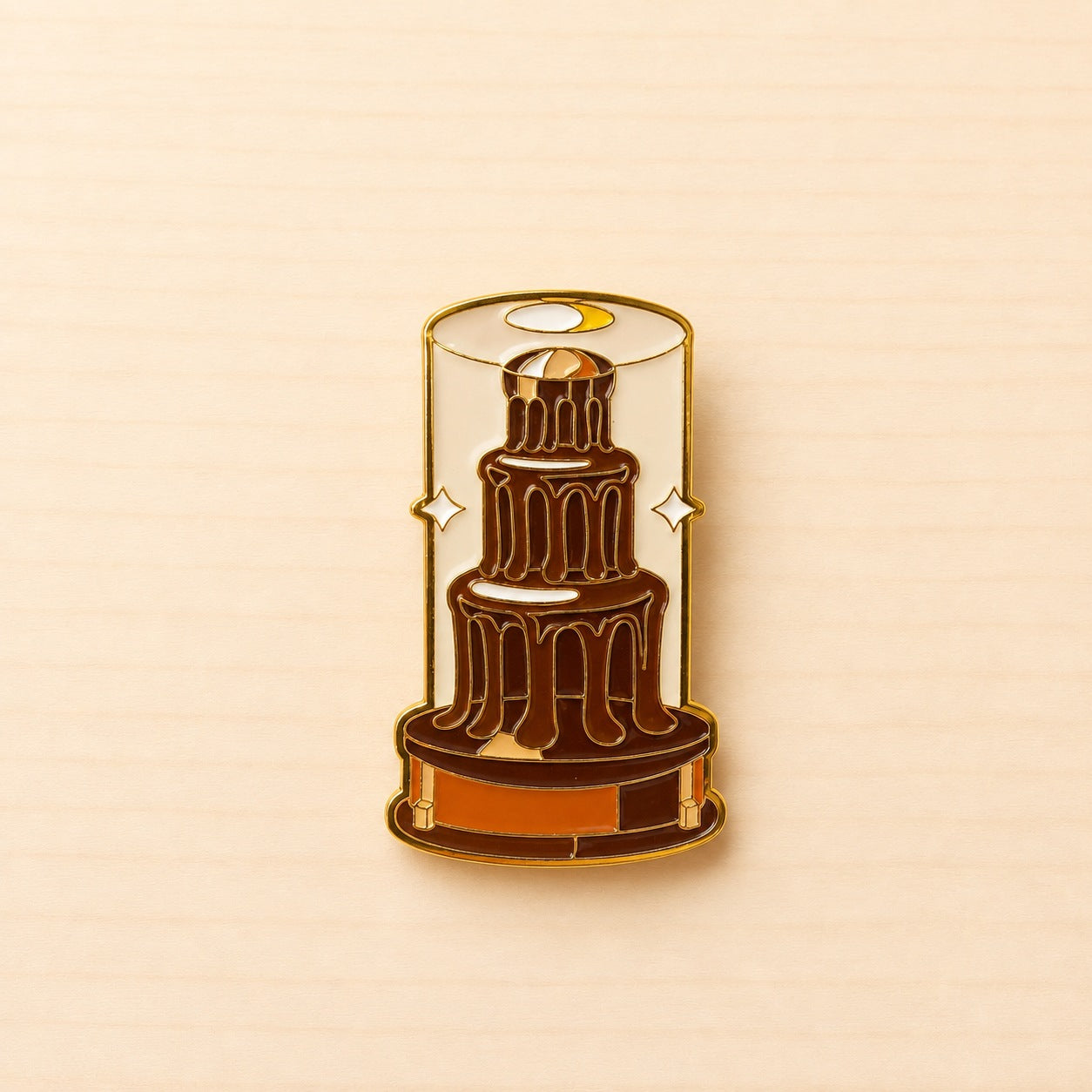 Metall-Ansteck-Pin mit mehrstöckigem braunem Schokobrunnen in einer hellen Vitrine und goldfarbenen Konturen.