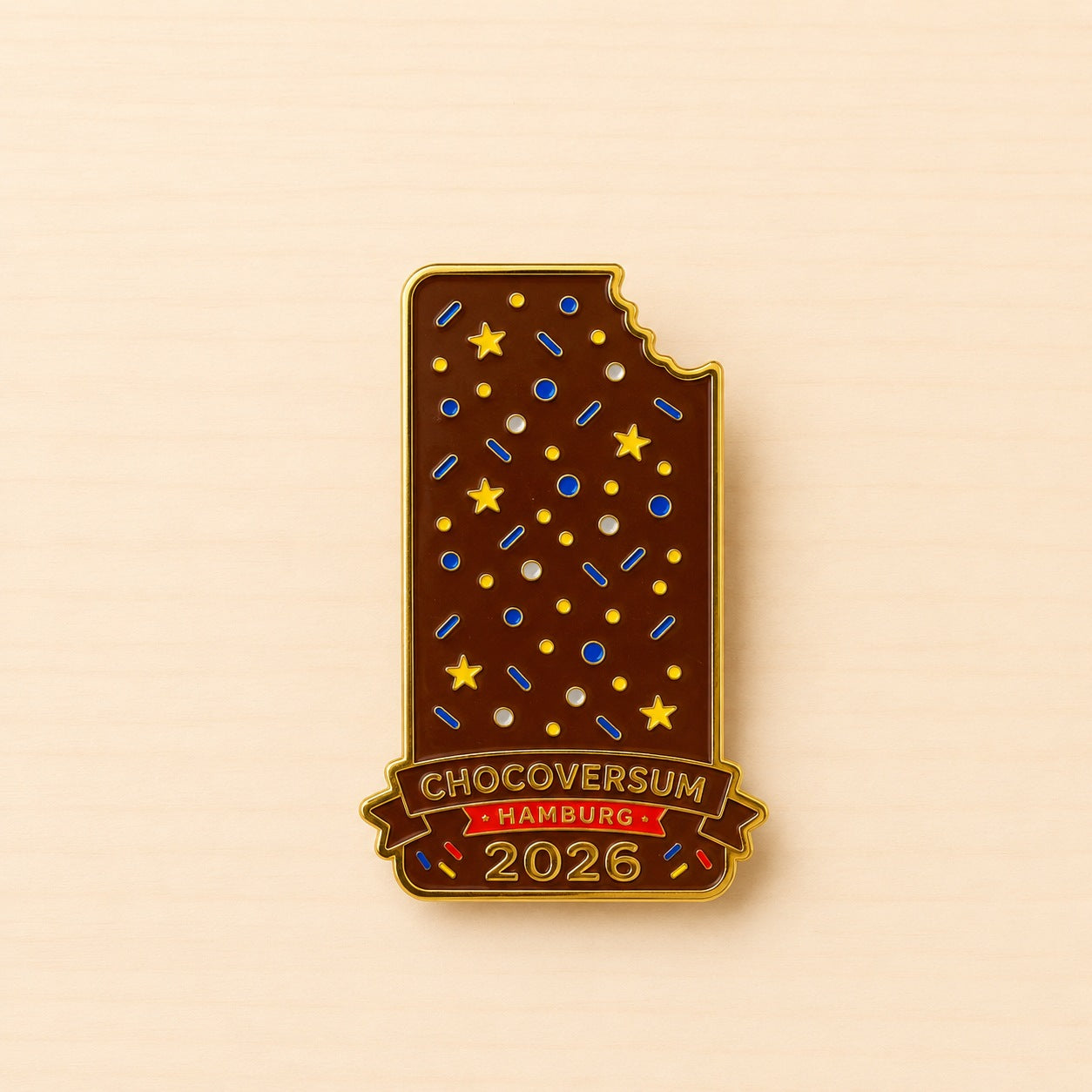 Metall-Ansteck-Pin 2026 als braune Schokoladentafel mit angebissener Ecke, bunten Streuseln und Schriftzug „CHOCOVERSUM Hamburg 2026“.