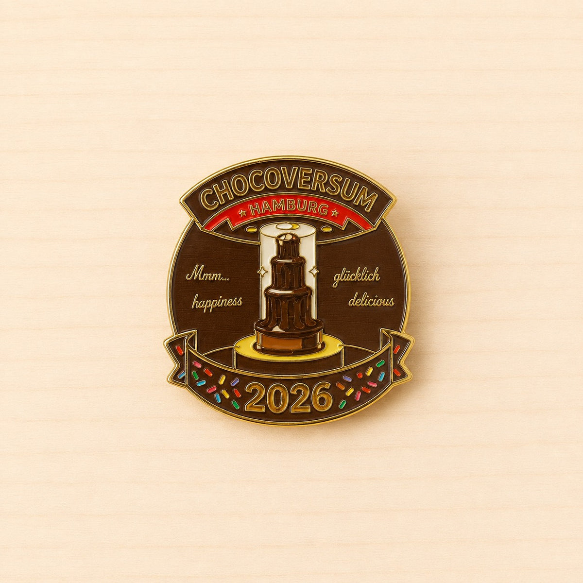 Runder Metall-Ansteck-Pin 2026 mit mehrstöckigem Schokobrunnen, Schriftzug „CHOCOVERSUM Hamburg“ und goldfarbenen Details.