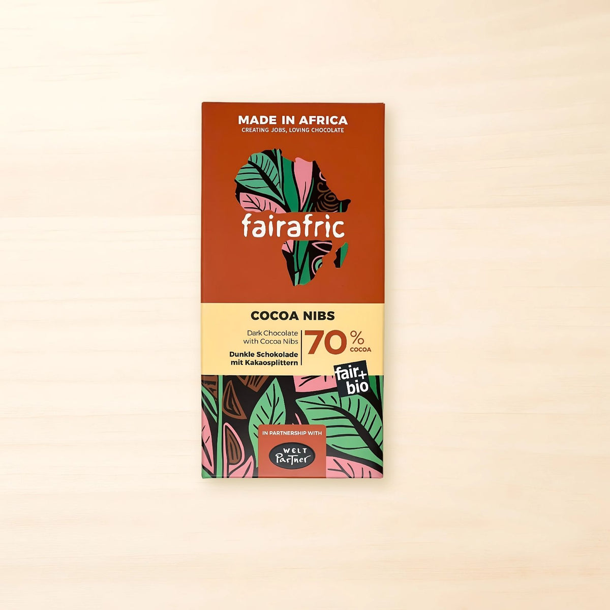 Orangefarbene fairafric Cocoa Nibs Schokoladentafel mit bunt gemusterter Afrika-Silhouette und floralen Motiven in Grün, Rosa und Braun. Auf der Vorderseite stehen „Cocoa Nibs“, „Dunkle Schokolade mit Kakaosplittern“ und „70 % Cocoa“ sowie der Hinweis auf die Partnerschaft mit WeltPartner.