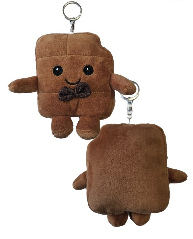 Brauner, quadratischer Plüsch-Schlüsselanhänger in Form einer Schokoladentafel mit lächelndem Gesicht und kleiner Fliege, liegend auf einer Holzoberfläche.