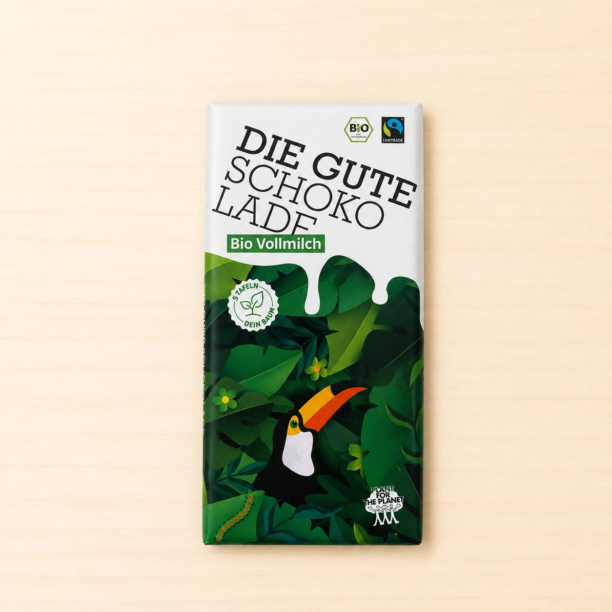 Verpackung der Schokoladentafel „Die Gute Schokolade – Bio Vollmilch“ mit weißem oberen Bereich, grüner Dschungelillustration mit Tukan im unteren Teil sowie BIO-, Fairtrade- und Plant-for-the-Planet-Logos. 