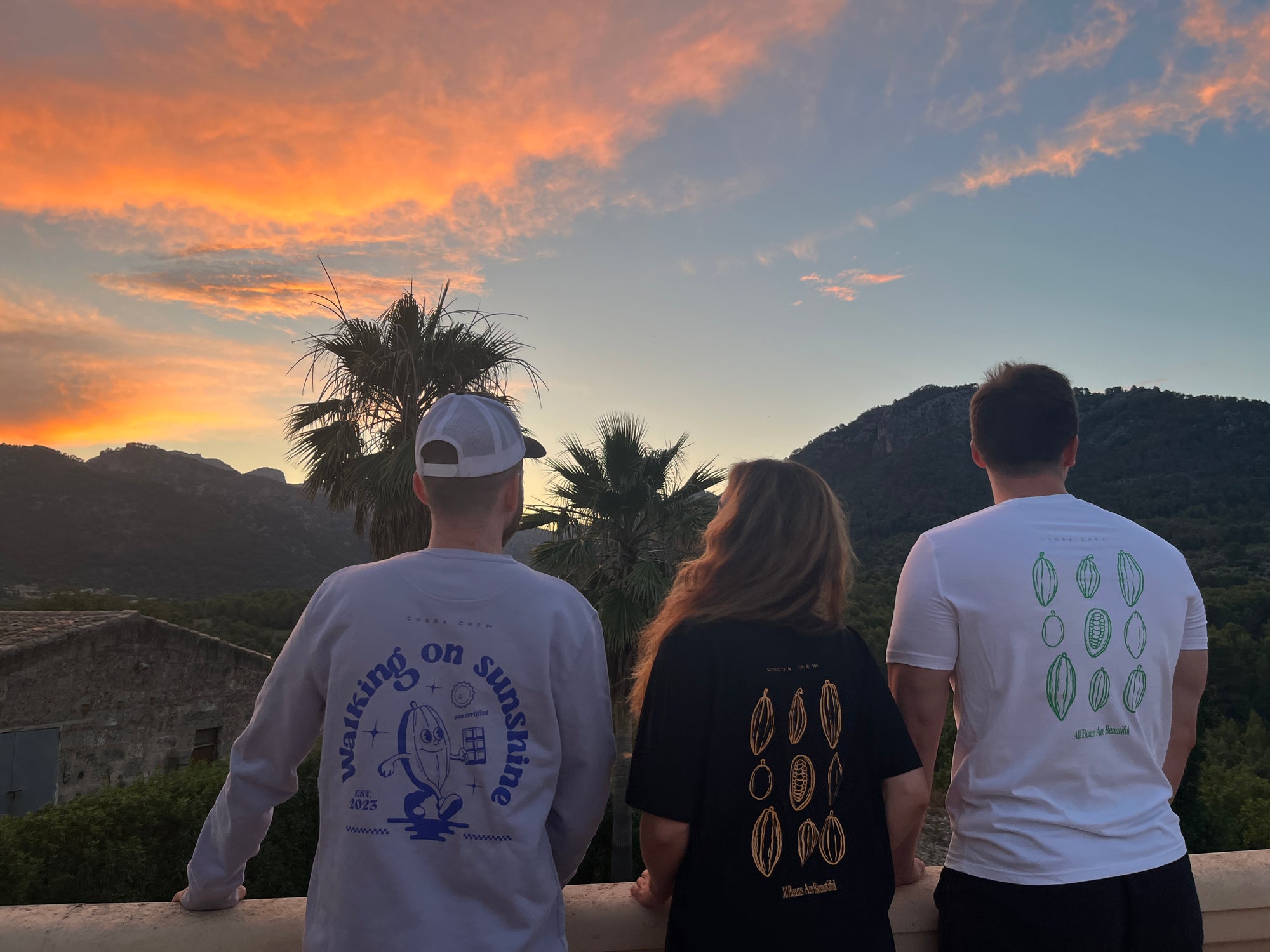 3 Personen stehen an einer Brüstung, im Hintergrund Palmen und Sonnenuntergang. Die Personen tragen Teile aus der neuen Chocoversum Merch-Linie.