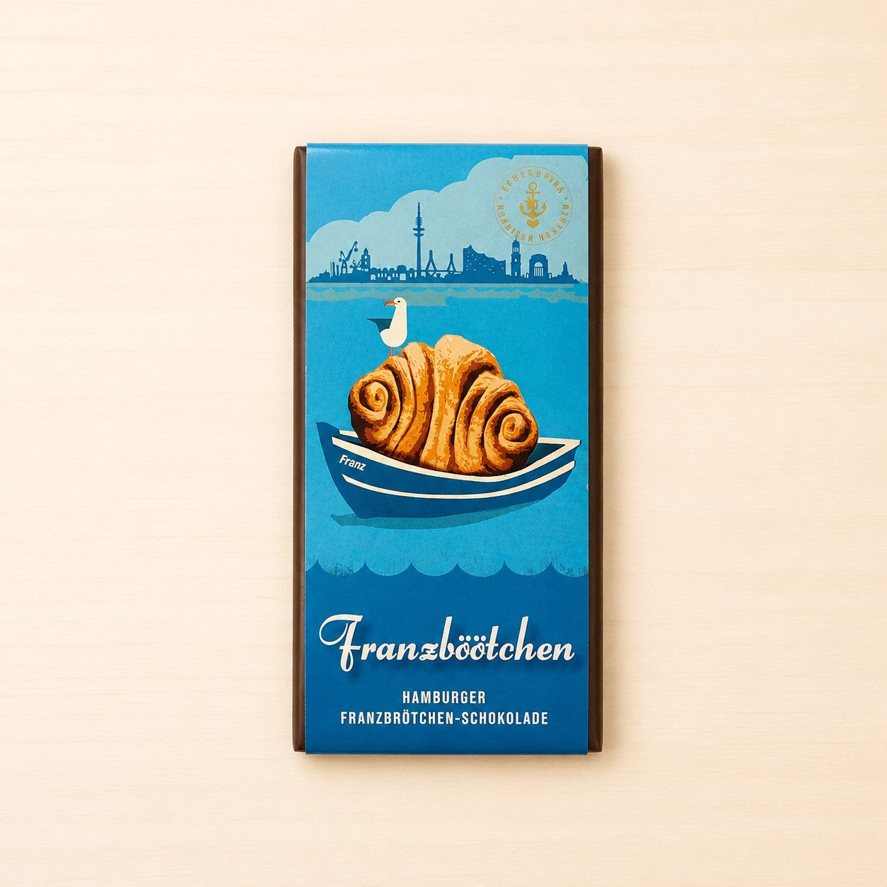 Hamburger Franzbrötchen-Schokolade in blauer Verpackung mit Illustration eines Franzbrötchens in einem kleinen Boot, einer Möwe und der Hamburger Skyline im Hintergrund. Aufdruck: „Franzbötchen – Hamburger Franzbrötchen-Schokolade“.